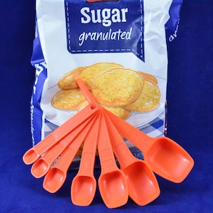 以下が含まれることがあります： グラニュー糖の袋の前に扇状に広げられた、オレンジ色の計量スプーン6本セット。袋は白と青のアクセントで、金色のクッキーのイラストが描かれています。スプーンには測定値が記載されています。
