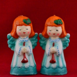 May include: Two ceramic angel figurines with red hair, blue dresses, and white wings. Each angel holds a red trumpet in their hands.