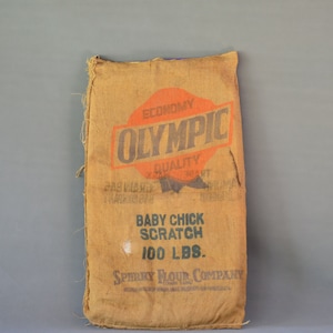 Könnte beinhalten: Ein brauner Jutesack mit dem Text "Economy Olympic Quality" und "Baby Chick Scratch 100 LBS." darauf gedruckt. Der Sack ist von der Sperry Flour Company.