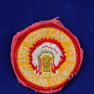 Puede incluir: Un parche bordado amarillo y rojo con el texto "East Carolina Council BSA Scout Camp". El parche presenta un tocado de nativo americano con un diseño de plumas rojas y blancas.