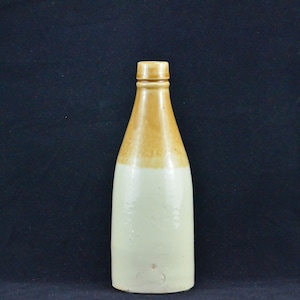 Puede incluir: Una botella de cerámica con la parte superior de color marrón claro y la inferior de color blanco. La botella tiene una forma redondeada y un labio ligeramente acampanado.