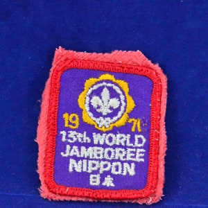 Puede incluir: Un parche vintage con un centro morado, borde rojo y detalles bordados en blanco y amarillo. El parche dice "1971 13th WORLD JAMBOREE NIPPON" con un emblema de flor de lis. También se ven caracteres japoneses.