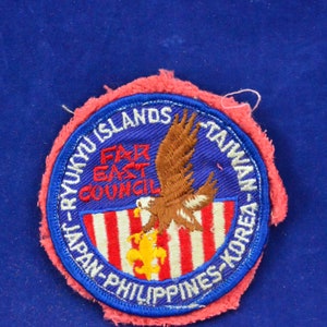 May include: A blue and white embroidered patch with the text "Far East Council" and the names of countries including Japan, Philippines, Taiwan, Korea, and Ryukyu Islands. A brown eagle with a yellow fleur-de-lis is in the centre of the patch.