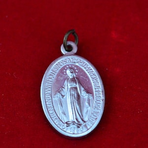 Op de afbeelding: Ovale, zilverkleurige hanger met een religieuze afbeelding van een vrouw met uitgestrekte armen. De hanger heeft een lus en is voorzien van tekst. De achtergrond is dieprood.