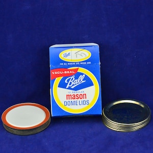 Peut inclure: Une boîte bleue et jaune de 12 couvercles de bocaux Mason réguliers de la marque Ball. La boîte est sur une surface bleue avec deux piles de couvercles, un blanc et un doré.
