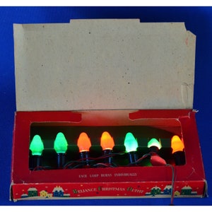 Könnte beinhalten: Eine Schachtel mit Vintage-Weihnachtslichtern mit roten, grünen und orangen Glühbirnen. Die Schachtel ist geöffnet und die Lichter sind sichtbar. Auf der Schachtel steht "Reliance Christmas Outfit" und "Each lamp burns individually".