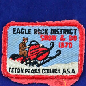 Op de afbeelding: Een blauw en wit geborduurde patch met een rode sneeuwscooter en een persoon die erop rijdt. De tekst op de patch luidt "Eagle Rock District Show & Do 1970 Teton Peaks Council, B.S.A."