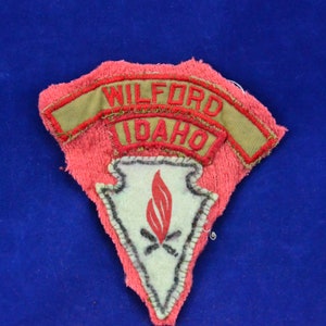 Puede incluir: Un parche bordado de color rojo, blanco y verde con el texto "WILFORD IDAHO" en rojo. El parche tiene forma de triángulo con una punta puntiaguda y una base curva. El parche tiene una punta de flecha blanca con una llama roja y dos palos cruzados en el centro.
