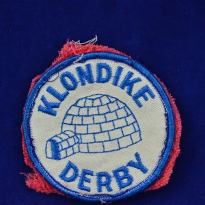 Puede incluir: Parche redondo con las palabras "KLONDIKE DERBY" en letras azules. El parche presenta un diseño de iglú azul sobre un fondo crema, con un borde rojo y azul.