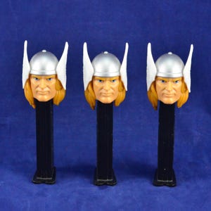 Puede incluir: Tres dispensadores PEZ de Thor. Cada dispensador presenta un casco plateado, cabello rubio y una expresión severa. Los dispensadores tienen una base negra y están sobre un fondo azul. Un artículo de colección para los fanáticos del superhéroe de Marvel.