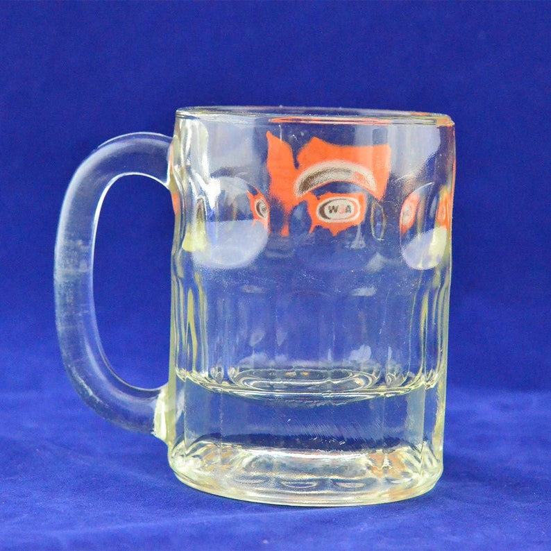 May include: A clear glass mug with a handle. The mug has a ribbed design and a logo that says "WA" in a circle. The logo is orange and white.