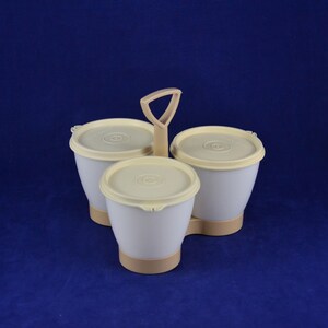 May include: A three-part serving dish with a handle. The set includes three white containers with cream-colored lids and bases. The handle is also cream-colored. The containers are arranged in a triangular shape.