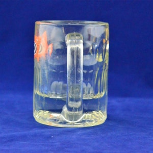 May include: A clear glass mug with a handle. The mug has a design of a fish with orange and black details.