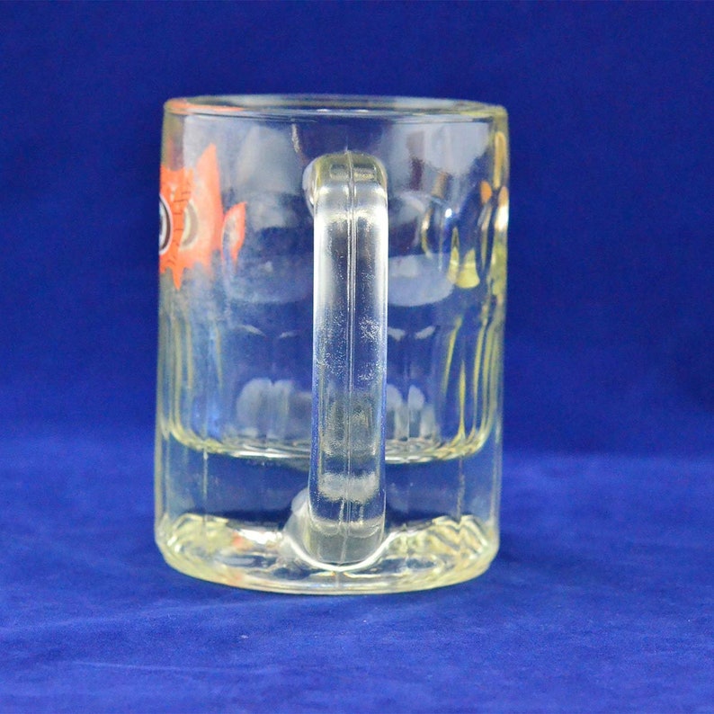 May include: A clear glass mug with a handle. The mug has a design of a fish with orange and black details.