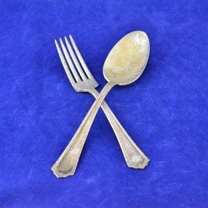 May include: A silver fork and spoon crossed against a blue background. The fork has four tines, and the spoon has a rounded bowl. Both utensils have decorative edges and appear to be vintage.