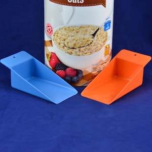May include: Two plastic scoops, one blue and one orange, are positioned in front of a container of oats. The container features an image of oatmeal with berries. The scoops are angled, with a small hole at the top.