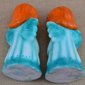 May include: Two ceramic angel figurines with orange hair and blue wings. The angels are standing on a white surface.