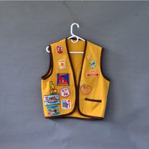 May include: A yellow vest with brown trim and multiple patches. The patches feature various designs, including state emblems and text. The vest is hanging on a white hanger against a gray background. The vest has a pocket on the right side.