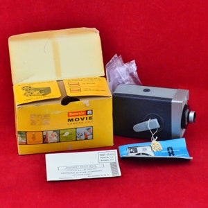 May include: A vintage Brownie 8 Movie Camera set. The set includes a yellow box with the camera name, a black and silver camera, a blue and white camera accessory, and a white envelope. The box is open, revealing the camera and accessories.