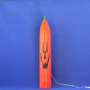 Può includere: Una decorazione alta e arancione a forma di zucca di Halloween con un volto sorridente intagliato. La zucca ha un gambo verde e un cavo di alimentazione bianco. Lo sfondo è blu.