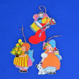 May include: Three vintage Christmas ornaments featuring illustrations of girls. One ornament depicts a girl carrying a Christmas tree, another shows a girl with a dog and a doll, and the last one shows a girl in a stocking with a drum and a toy.