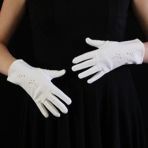 Op de afbeelding: Een paar witte handschoenen met decoratieve uitgesneden bloempatronen. De handschoenen zijn gemaakt van een gladde stof en hebben een klassiek ontwerp. De handschoenen worden tegen een zwarte achtergrond getoond.