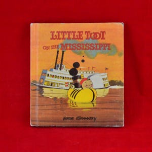Op de afbeelding: Vintage kinderboek getiteld "Little Toot on the Mississippi" door Hardie Gramatky. De omslag toont een illustratie van een raderstoomboot en een gele sleepboot met een lachend personage. De achtergrond is een zonsondergang.