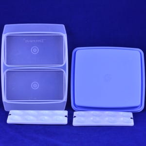 May include: A clear plastic food storage container with two compartments and a blue lid. Two white serving trays are at the bottom. The container is on a blue background.