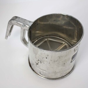 May include: A vintage, stainless steel flour sifter with a handle. The sifter has a cylindrical body with a mesh screen at the bottom. The handle is attached to the side of the sifter. The sifter is designed for baking and cooking.