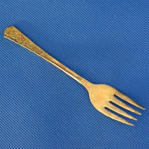 May include: A gold-toned fork with a floral design on the handle. The fork is on a blue textured surface.