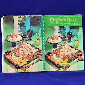 May include: A vintage cookbook titled "Our Favorite Salads" with a green cover and a photo of a salad platter with chicken, tomatoes, cucumbers, olives, and eggs. The book features 2000 recipes from home economics teachers.
