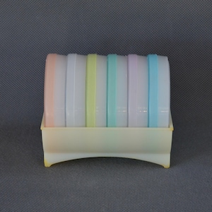 Puede incluir: Un conjunto de recipientes de plástico de colores pastel con tapas, dispuestos en un soporte rectangular. Las tapas son blancas con bandas rosas, amarillas, verdes, azules y moradas. El soporte es de color amarillo pálido.