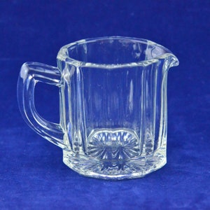 Puede incluir: Una jarra de crema de vidrio transparente y facetado con asa y boquilla. La jarra tiene forma rectangular con líneas verticales y un diseño decorativo en forma de estrella en la base. El fondo es azul liso.