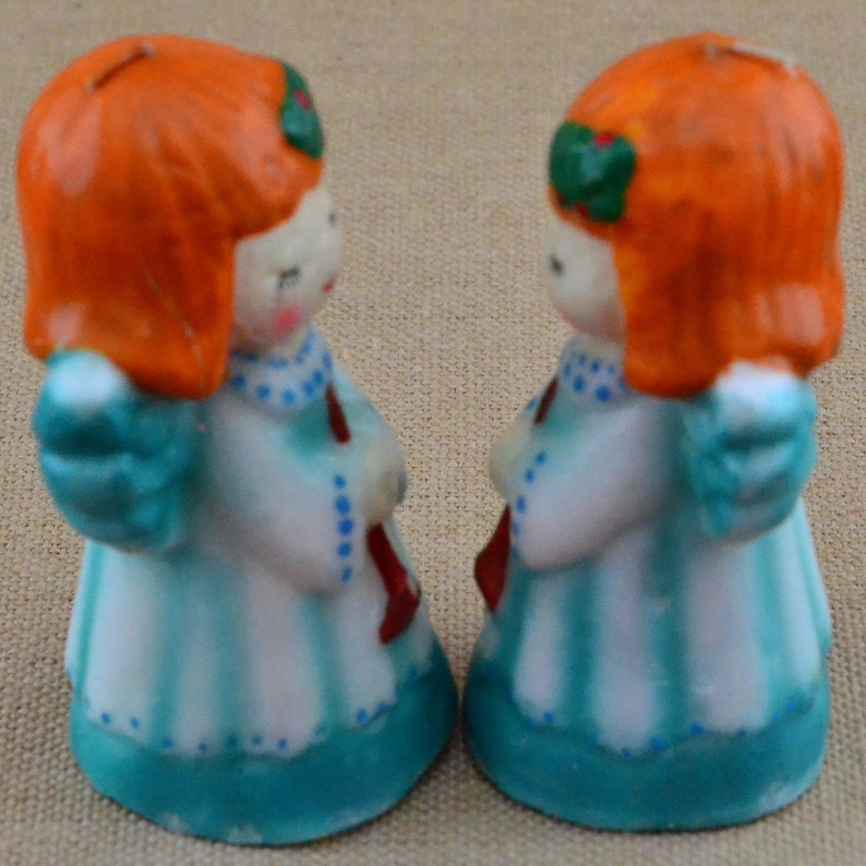 May include: Two small, white porcelain angel figurines with red and green accents. The angels have orange hair and blue and white dresses. They are facing each other.