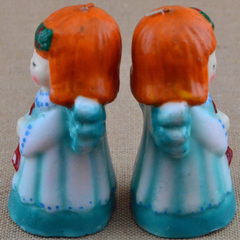 May include: Two red-haired angel candle figurines with blue and white dresses and wings. The candles are standing on a light brown surface.