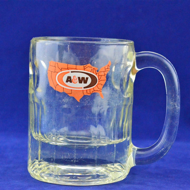 May include: A clear glass mug with a handle. The mug has a raised design and a sticker on the front that says "A&W" and features an outline of the United States.