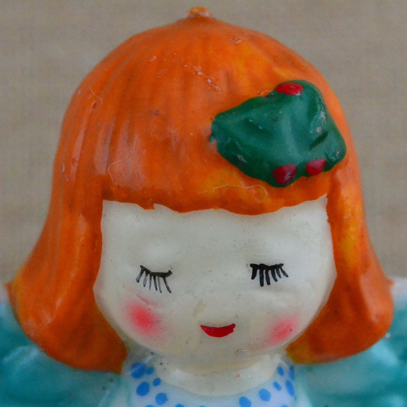May include: A ceramic figurine of a girl with orange hair, closed eyes, and a green holly leaf with red berries on her head. She is wearing a blue dress with white polka dots.