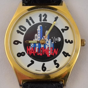 May include: A gold-toned wristwatch with a black leather band. The watch face features a white background with black numerals and a central graphic of a Halloween scene with the word "HALLOWEEN" in red. The watch hands are gold and green.
