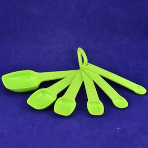 Puede incluir: Un juego de seis cucharas medidoras de color verde lima de diferentes tamaños, conectadas por un anillo. Las cucharas son de plástico y están dispuestas sobre un fondo azul. Cada cuchara tiene marcas de medición.