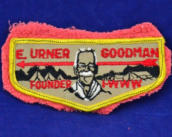 May include: A tan embroidered patch with a red arrow, a cartoon image of a man with a mustache, and the text "E. Urner Goodman Founder H.W.W.W."