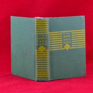 Puede incluir: Un libro de tapa dura verde titulado "Girl Scout Hand Book" con letras doradas y rayas en el lomo. El libro está cerrado y sobre una superficie roja.