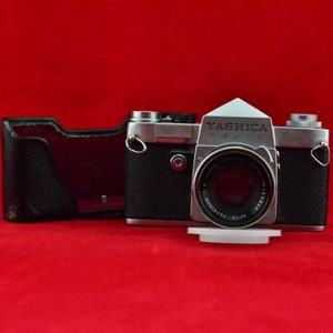 May include: A silver YASHICA PENTAX camera with a black leather case. The camera has a silver lens with the text "YASHICA AUTO 1:1.8 50mm" printed on it.