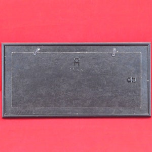 Pode incluir: Parte traseira de uma moldura retangular preta com uma superf&iacute;cie texturizada. A moldura tem dois ganchos de metal para pendurar e um suporte central para montagem. O fundo &eacute; vermelho liso.
