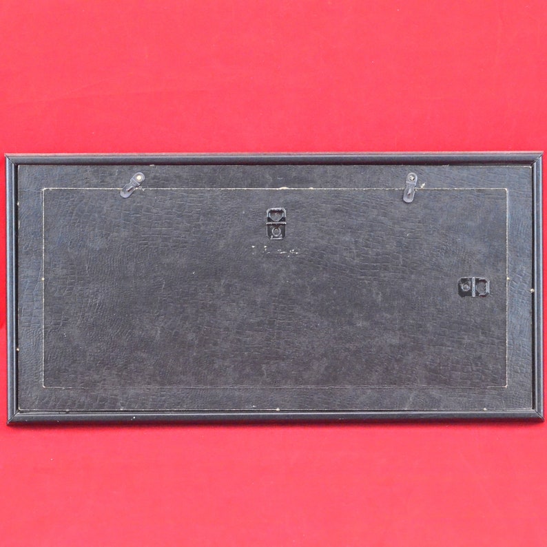 Pode incluir: Parte traseira de uma moldura retangular preta com uma superf&iacute;cie texturizada. A moldura tem dois ganchos de metal para pendurar e um suporte central para montagem. O fundo &eacute; vermelho liso.