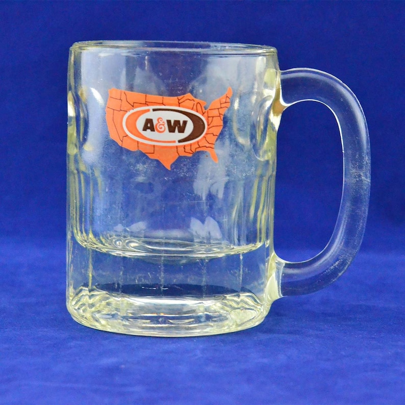 May include: A clear glass mug with a handle. The mug has a raised design and a sticker on the front that says "A&W" and features a map of the United States.