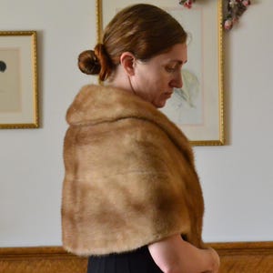 May include: A light brown fur stole with a soft, dense texture and a wide collar that drapes over the shoulders. The garment features a smooth, uniform appearance with a short pile, providing full coverage across the upper back and chest area.