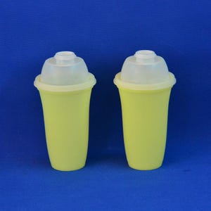 May include: Two identical yellow plastic cups with translucent white lids. Each cup features a tapered cylindrical body and a smooth, rounded rim where the lid attaches. The lids are dome-shaped with a small circular opening at the very top center.