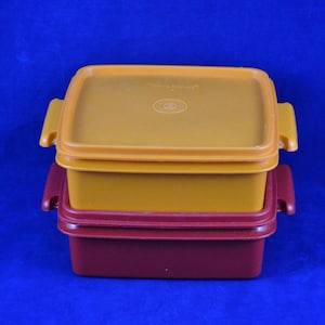 Op de afbeelding: Drie rechthoekige voedselcontainers op elkaar gestapeld. De bovenste container is goudgeel, de middelste is donkerder geel en de onderste is dieprood. Elke container heeft een bijpassend deksel en zijhandgrepen.