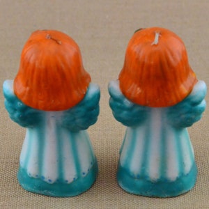 May include: Two small, teal and white angel candle holders with orange hair. The angels are facing away from each other.