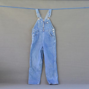 Könnte beinhalten: Hellblaue Jeans-Latzhose mit Frontreißverschluss, verstellbaren Trägern und mehreren Taschen. Die Latzhose hängt an einer blauen Wäscheleine vor grauem Hintergrund. Das Kleidungsstück hat Metallknöpfe und Nieten.